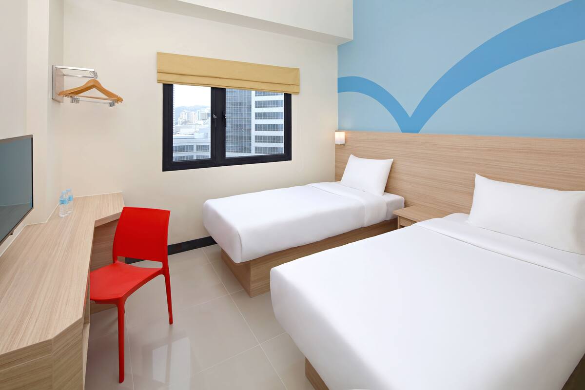 A hotel room with two beds, a desk, a red chair, a TV, and a window with a view of buildings.