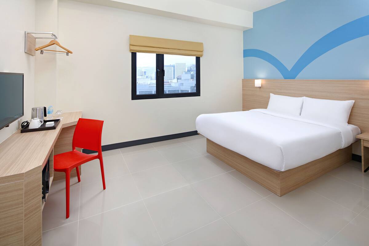 A hotel room with a bed, a desk with a chair, a TV, and a window with a view of buildings.