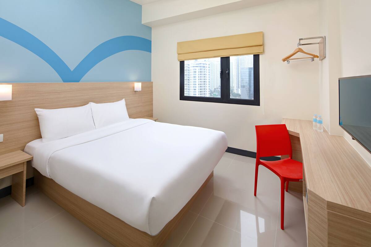 A hotel room with a large bed, a desk with a chair, a TV, a window with a view of buildings, and a wall with a blue design.