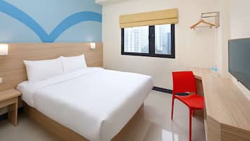 A hotel room with a large bed, a desk with a chair, a TV, a window with a view of buildings, and a wall with a blue design.