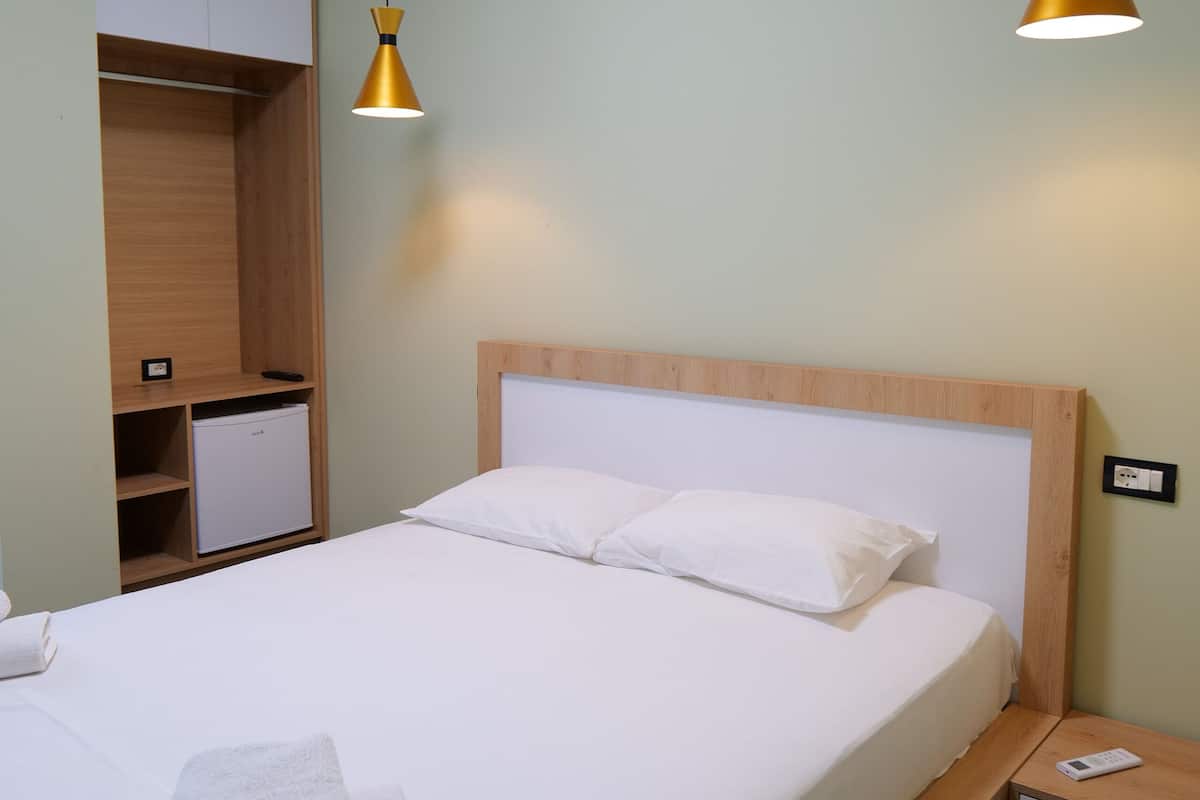 A neatly made bed with white linens and a folded towel, a wooden nightstand with a lamp, and a built-in wardrobe with a small refrigerator.