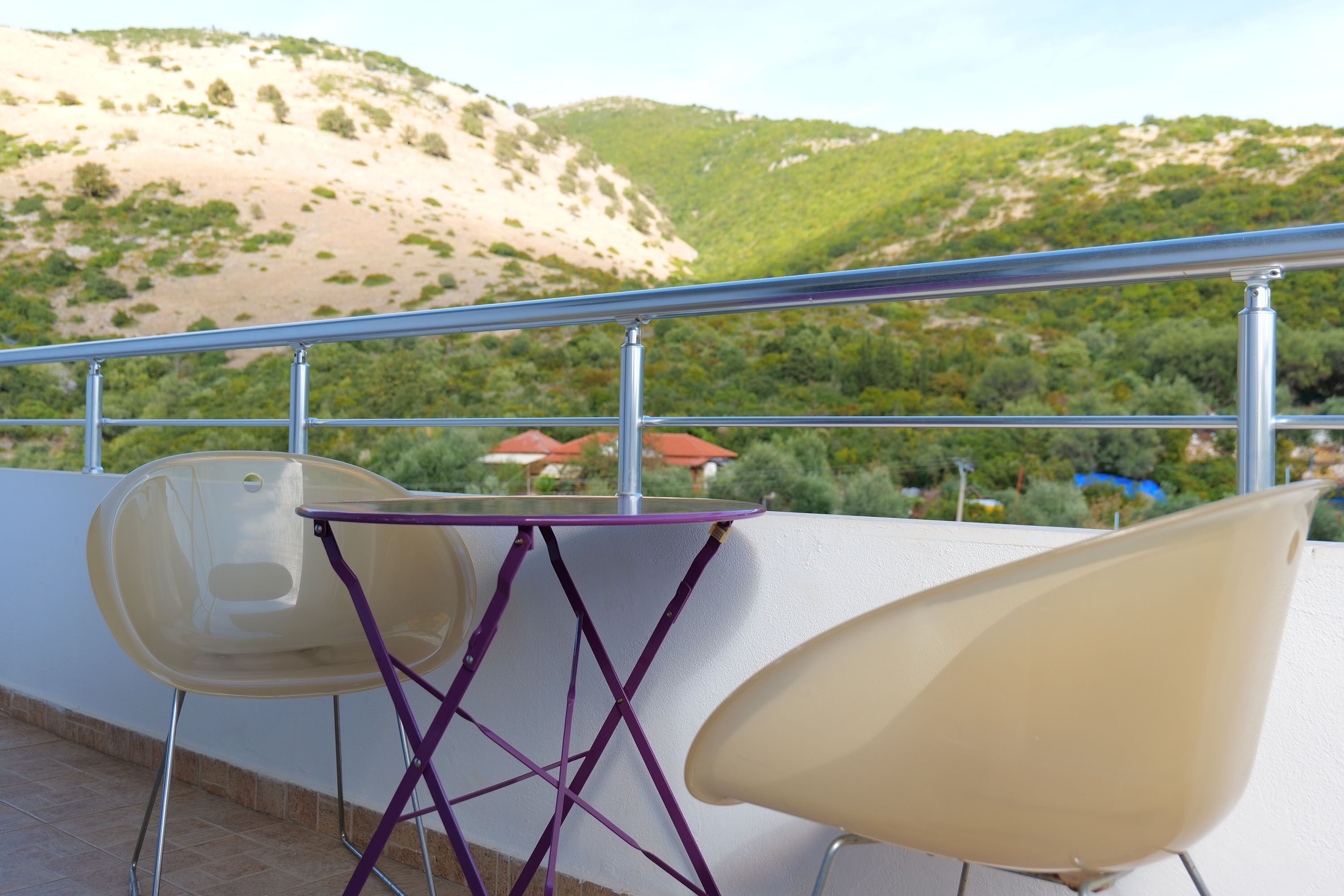 A balcony with two chairs and a table, overlooking a hilly landscape with greenery and distant buildings.