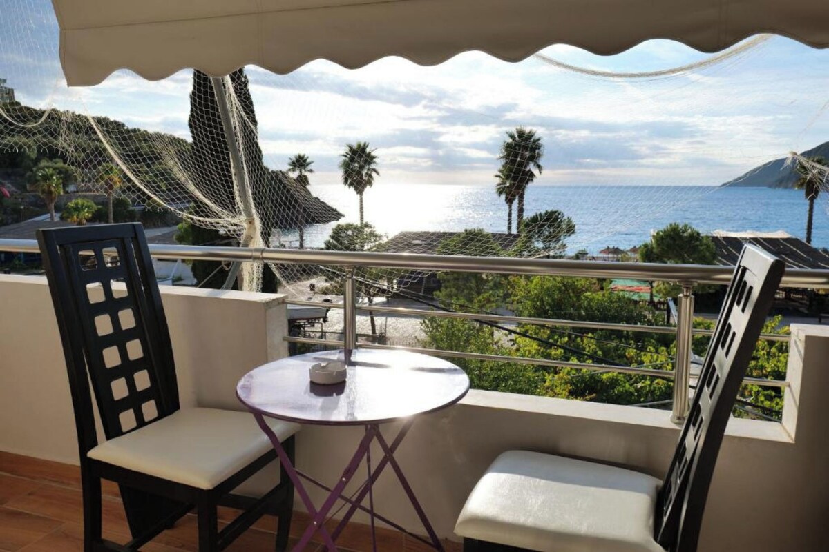 A balcony with a table and chairs, overlooking a coastal view with palm trees and a clear sky.