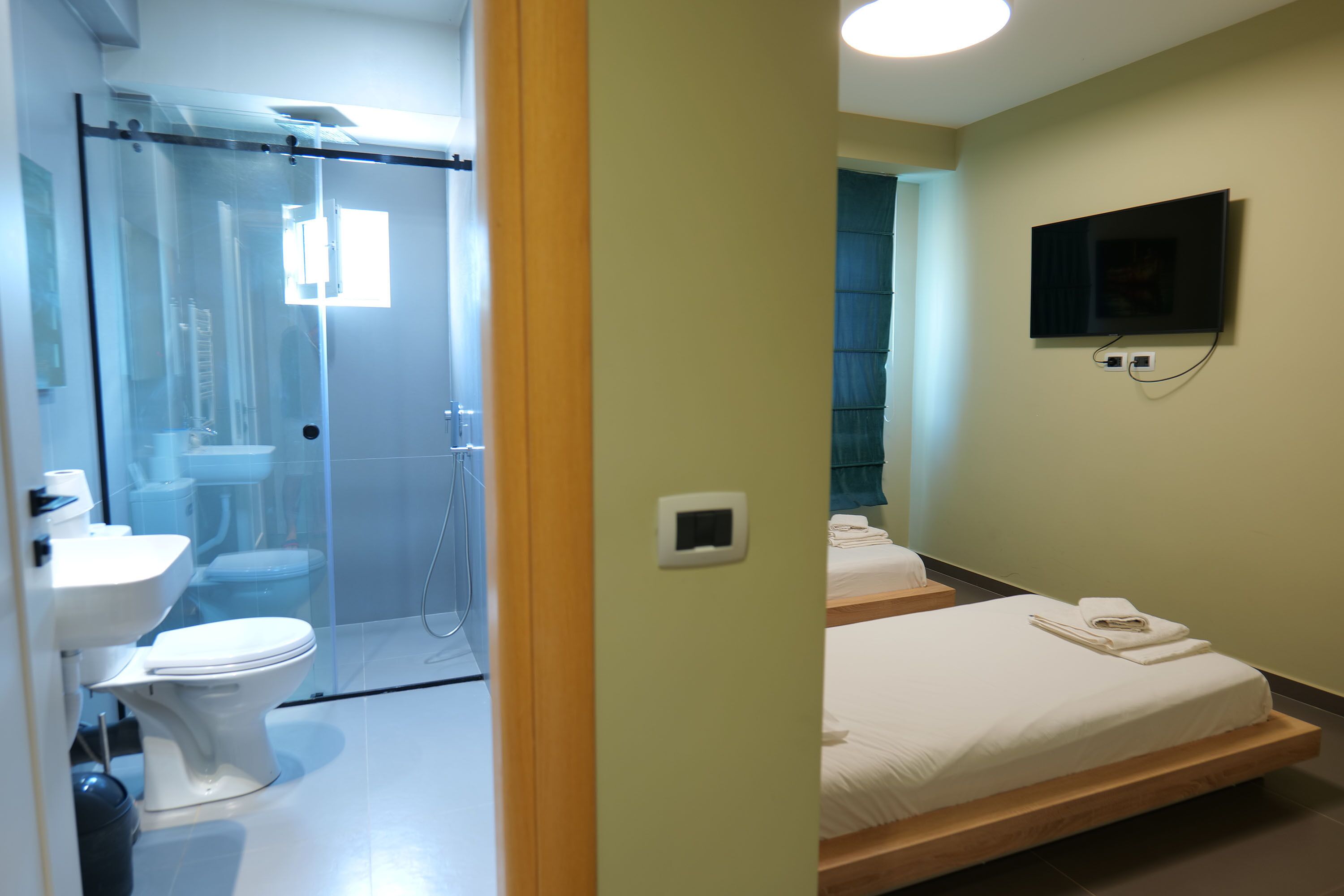 Habitación de hotel con cabina de ducha de vidrio, inodoro, lavabo y televisor de pantalla plana montado en la pared, además de dos camas con toallas.