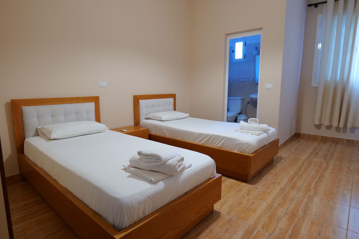 A hotel room with two single beds, wooden headboards, white linens, and a bathroom visible through an open door.