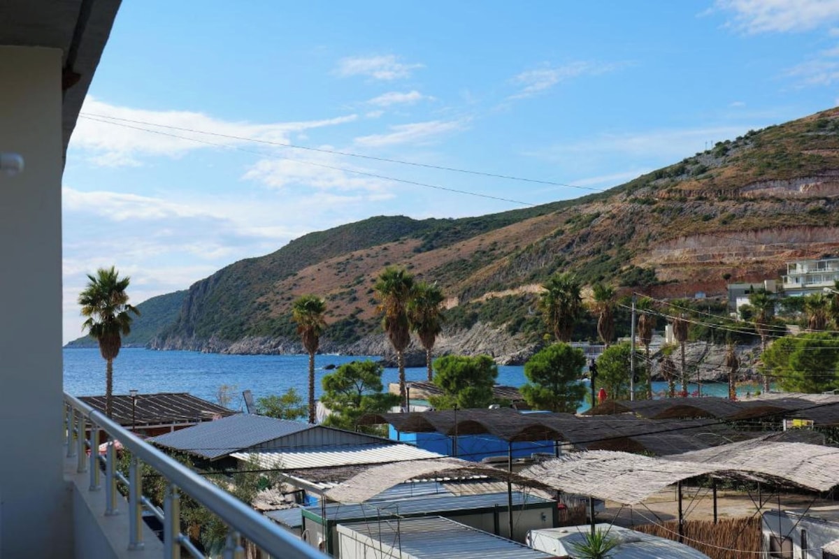 A coastal view with a balcony overlooking a beach, a hillside, and several buildings with flat roofs.