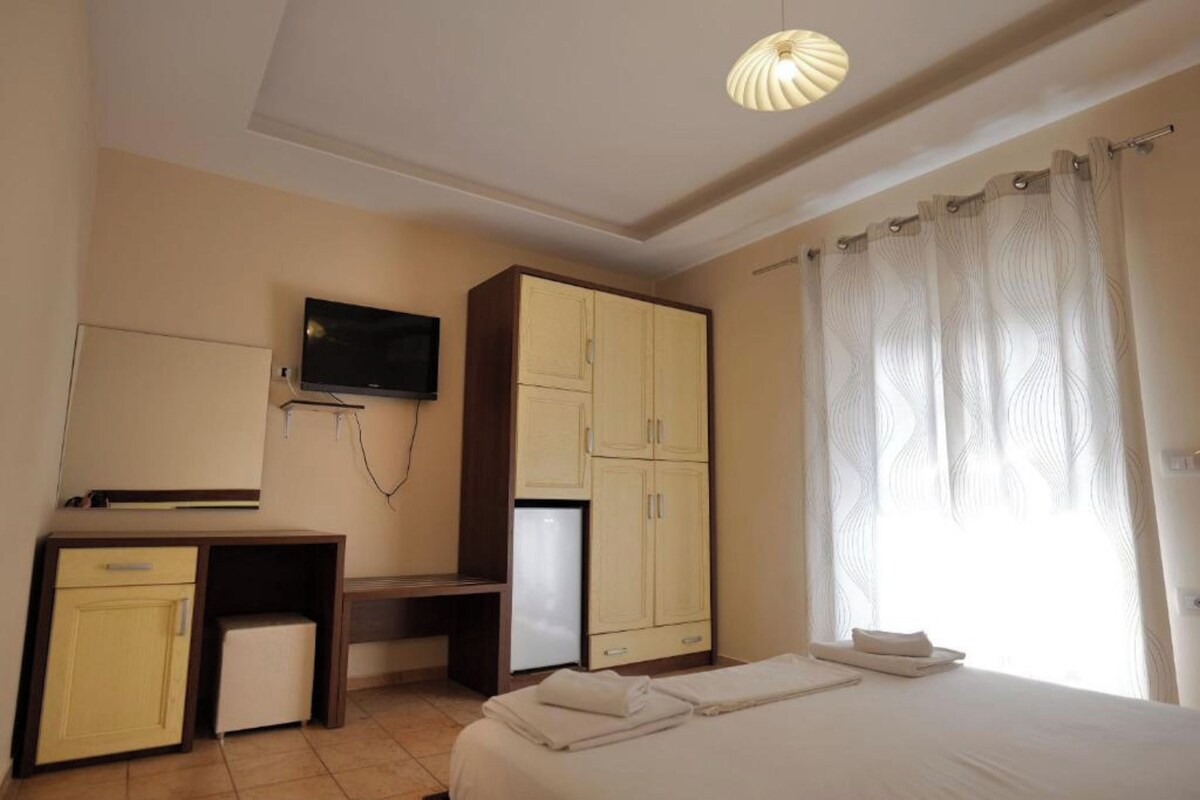 A hotel room with a bed, a desk with a television, a wardrobe, and a window with curtains.