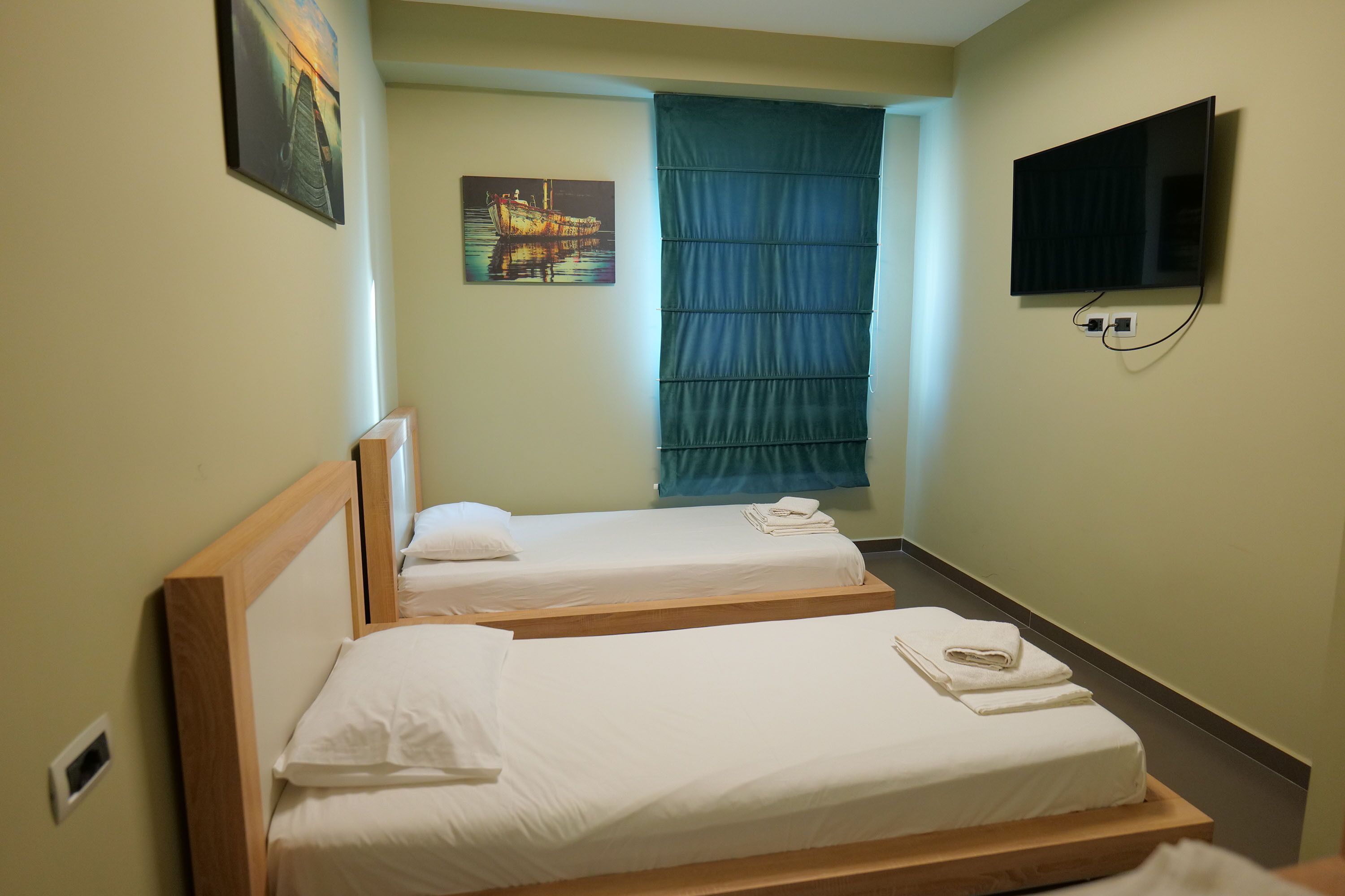A hotel room with two single beds, a television mounted on the wall, and a window with blue curtains.