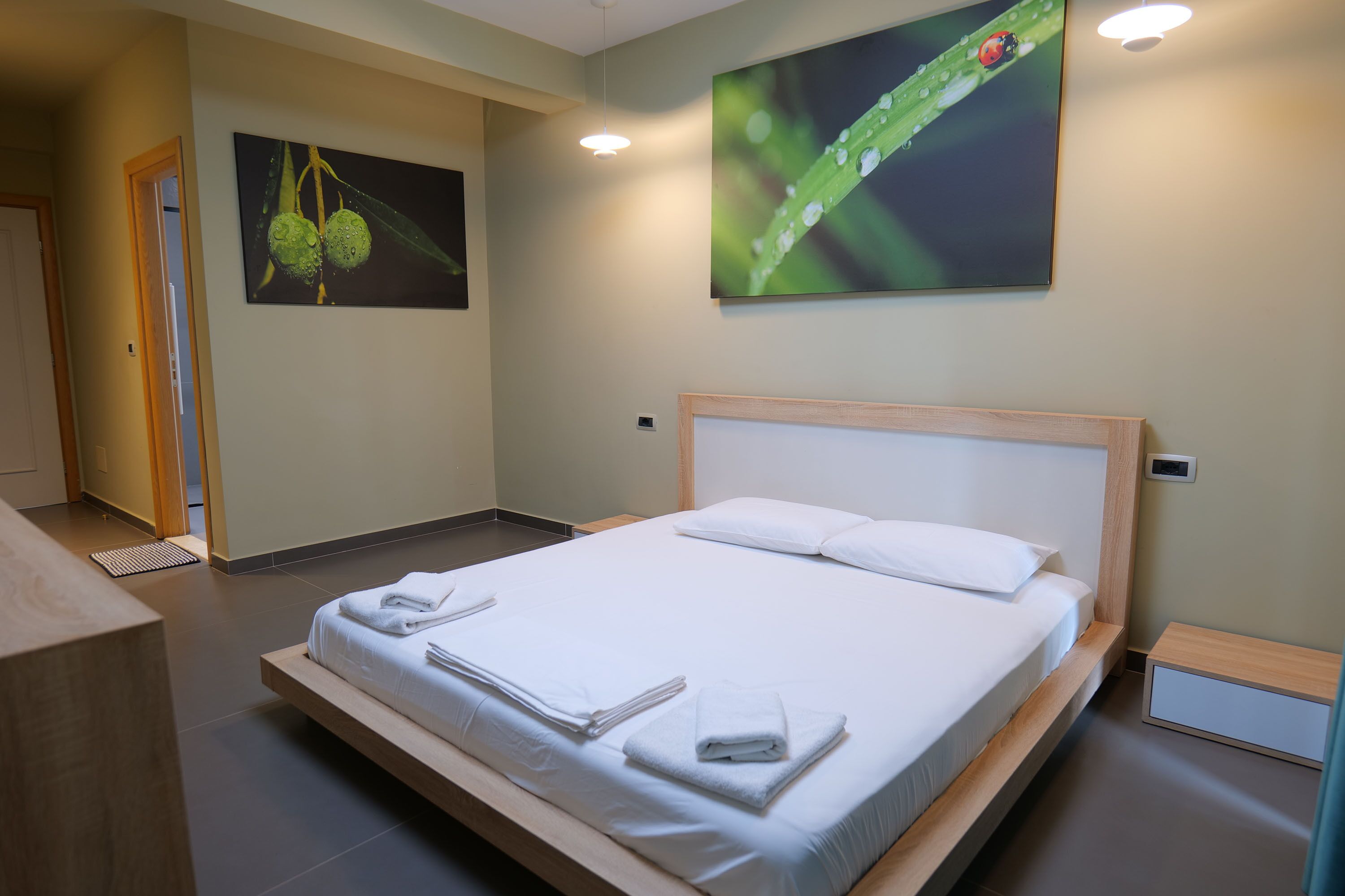 Una cama bien hecha con sábanas y toallas blancas, un cabecero de madera y un cuadro de una mariquita sobre una hoja en la pared.