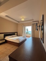 Habitación de hotel con cama, mesita de noche, un amplio ventanal con cortinas, una lámpara en el techo y vista al mar.