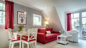 A modern living room with a red sofa, white chairs, a dining table, and a white armchair. There is a painting on the wall, a lamp, and a window with curtains. - Quietly situated apartment for up to 2 p. + infant, sauna, WLAN, balcony, bicycle shed (Prerow)