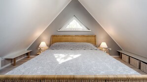 A bedroom with a sloped ceiling, a bed with a grey patterned bedspread, two bedside lamps, a hanging light fixture, and a window with a triangular design. - Quietly situated apartment for up to 2 p. + infant, sauna, WLAN, balcony, bicycle shed (Prerow)