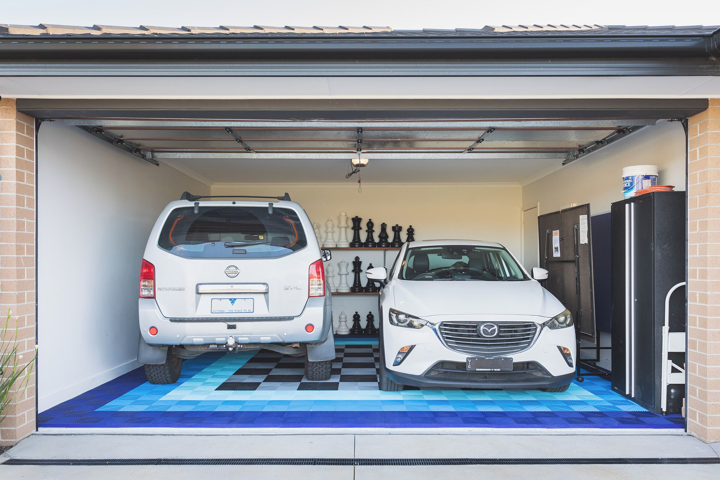 Un garage dans lequel sont garées deux voitures, un sol décoré comme un échiquier et un réfrigérateur placé à droite.