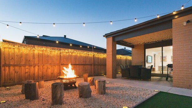A backyard with a fire pit surrounded by wooden stumps and gravel, string lights overhead, and a covered patio area with outdoor furniture.