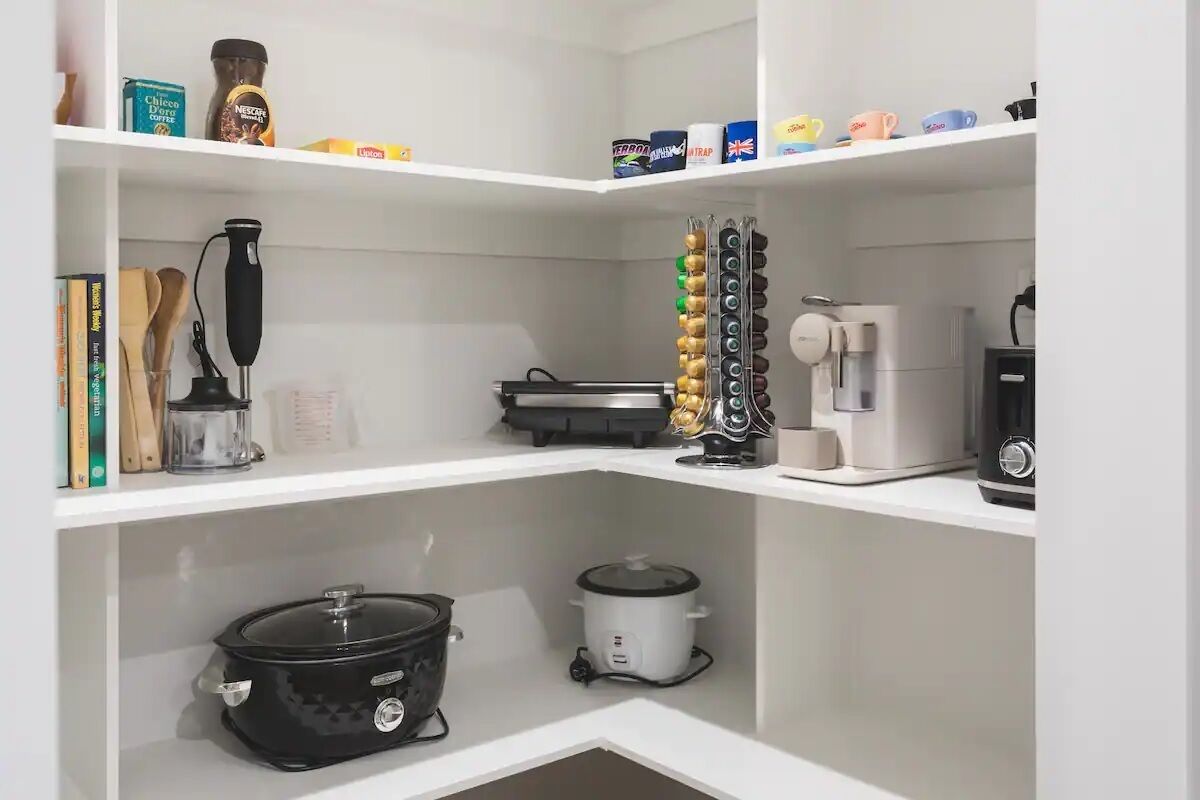 Een keuken met witte planken, een zwarte slowcooker, een witte slowcooker, een koffiezetapparaat en diverse keukengerei.