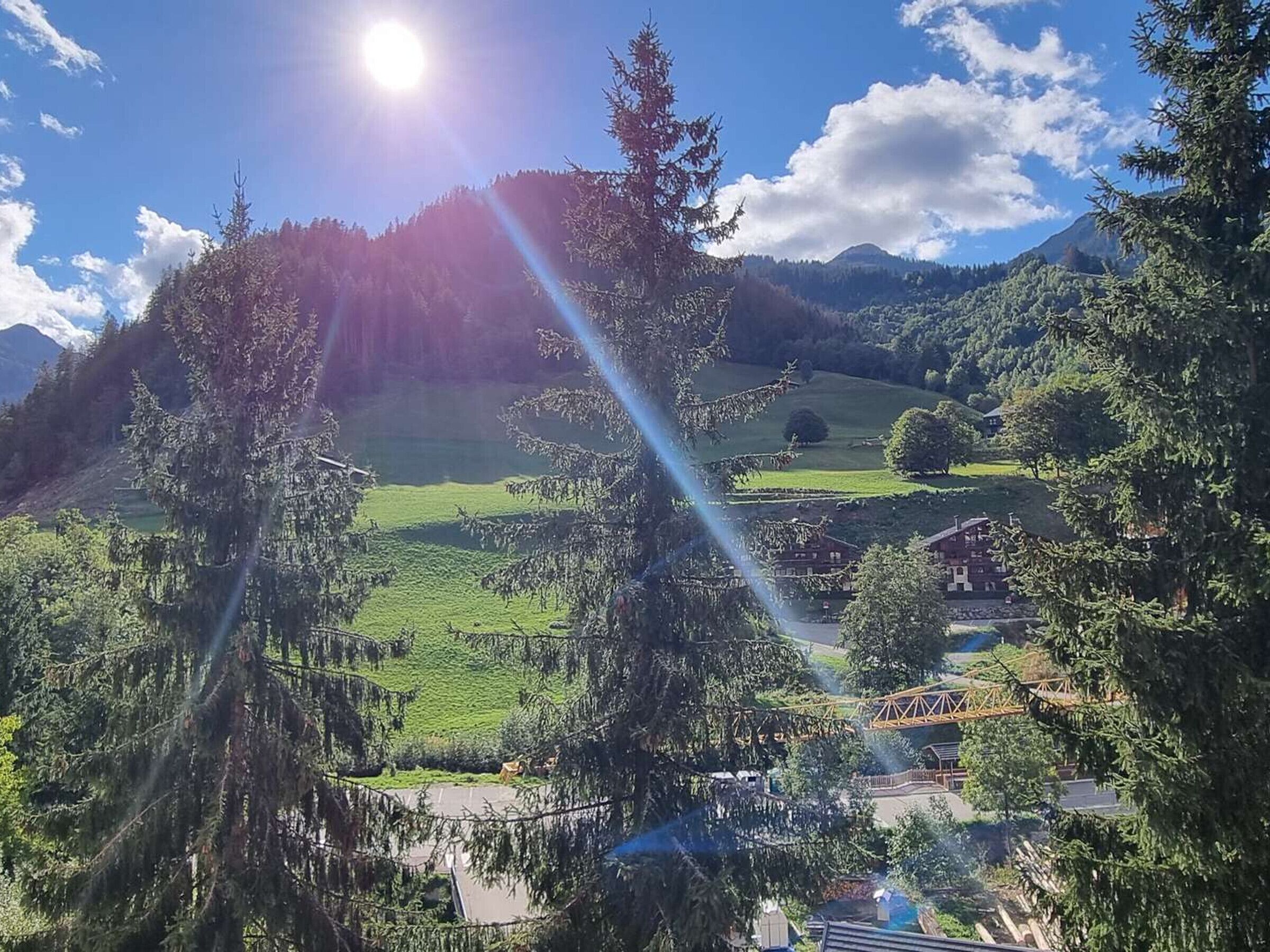 Eine gebirgige Landschaft mit blauem Himmel, der Sonne, die durch die Bäume scheint, und Blick auf ein Gebäude mit rotem Dach.