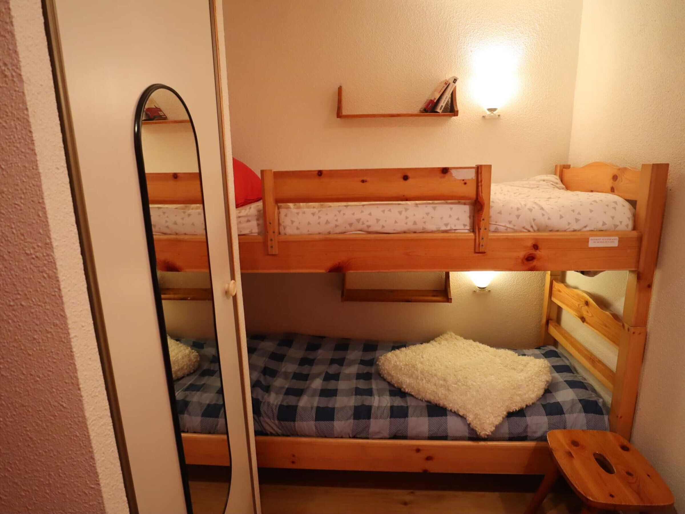A bunk bed room with a mirror, a shelf with books, and a small stool.