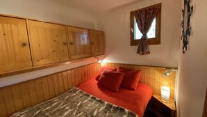 A bedroom with a wooden headboard, a bed with red bedding, a nightstand with a lamp, and a window with curtains. - Cozy apartment 3-5 pers, pool, near slopes, village center (Arêches-Beaufort)