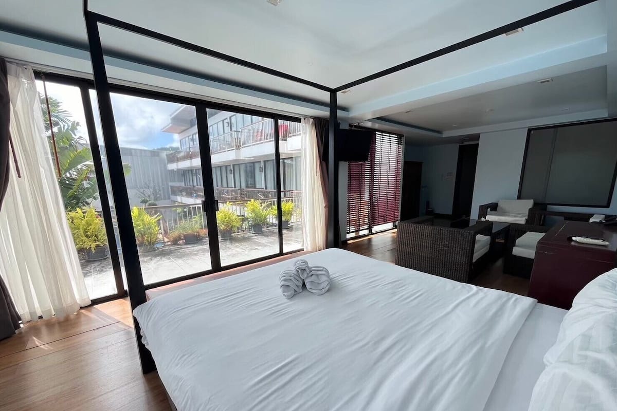 A modern hotel room with a large bed, a seating area with a sofa and armchairs, and a balcony with potted plants.