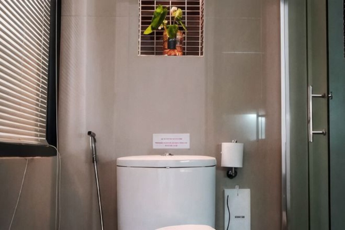 Une salle de bain comprenant des toilettes, une douche, un distributeur de serviettes et une petite fenêtre avec des plantes.