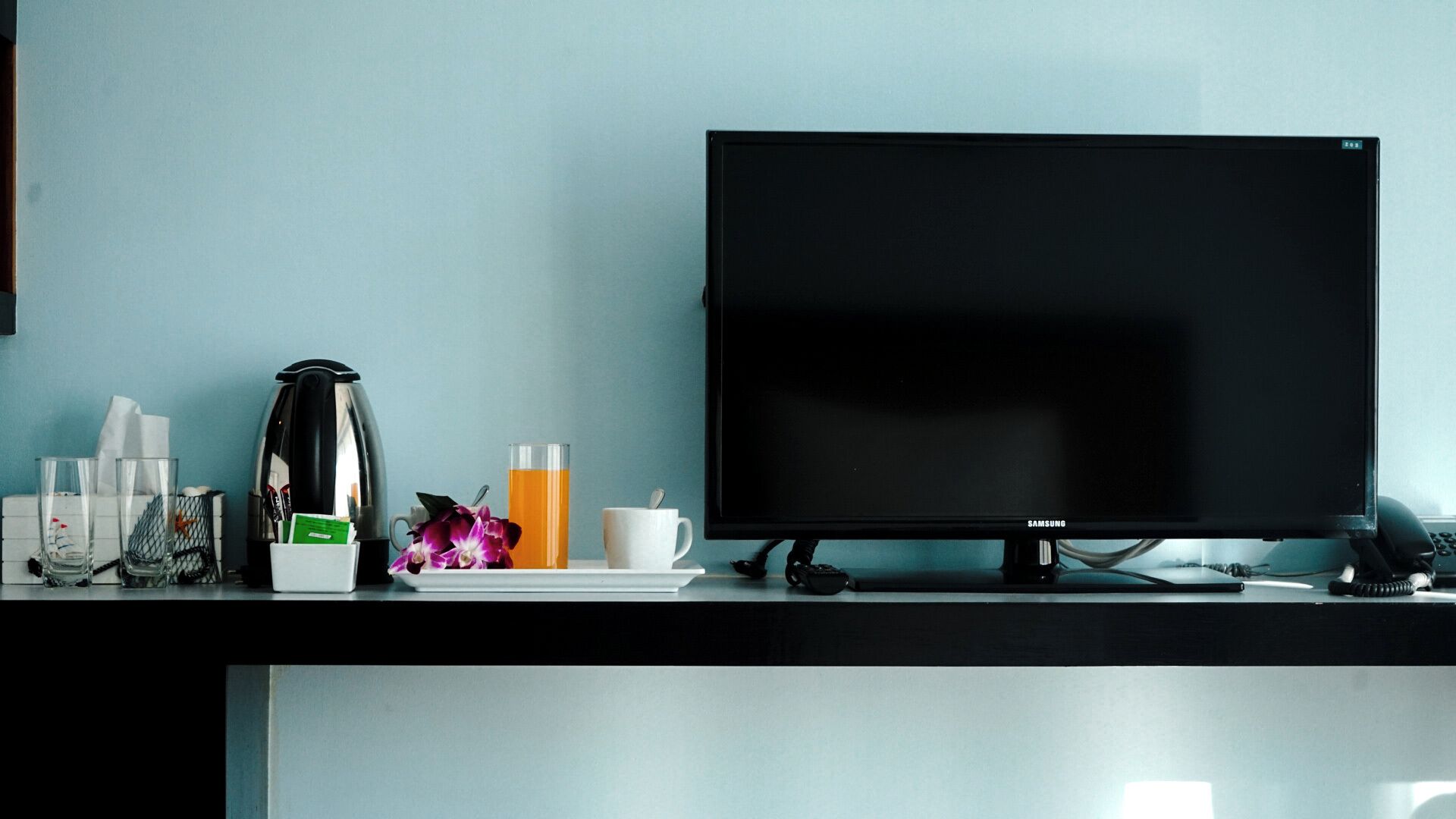 A modern hotel room with a black TV on a stand, a coffee maker, a glass of orange juice, a white mug, and a flower on the counter.
