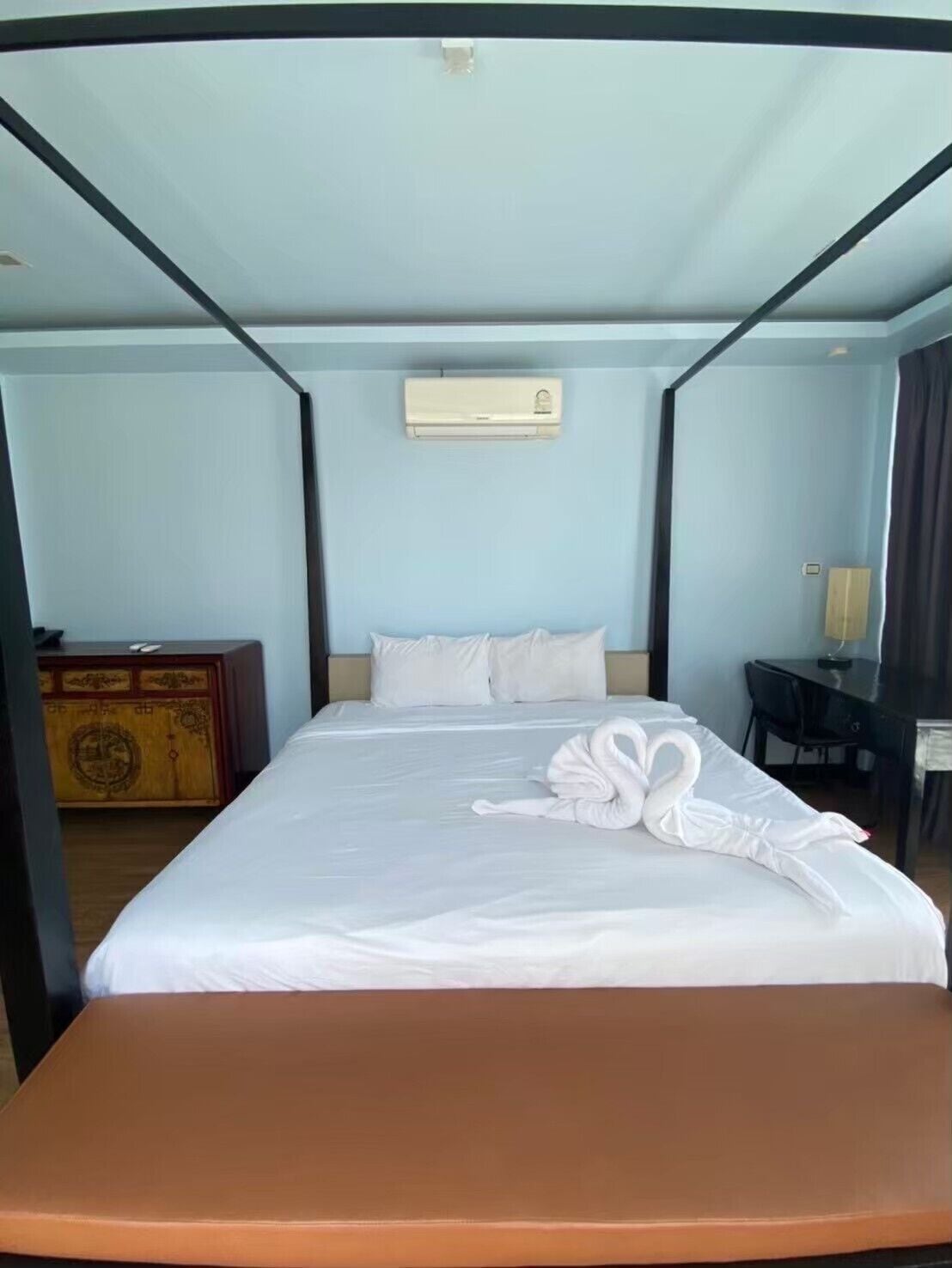 A hotel room with a bed, a desk, a chair, an air conditioning unit, and a wooden cabinet.