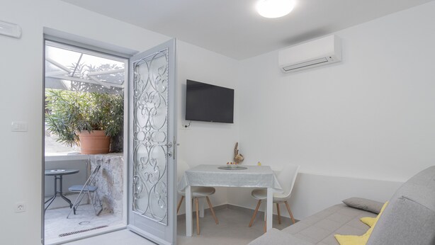 Una sala moderna con un sofá gris, una pequeña mesa con sillas, una planta en maceta y un televisor montado en la pared.