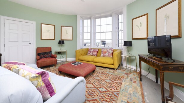 Een woonkamer met een gele bank, een rode fauteuil, een witte bank, een kamerkleed met patroon en ingelijste foto's aan de muren.