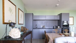 Een compacte keuken met grijze kastjes, een eettafel met stoelen en een klein kookeiland met een lamp en decoratieve objecten.