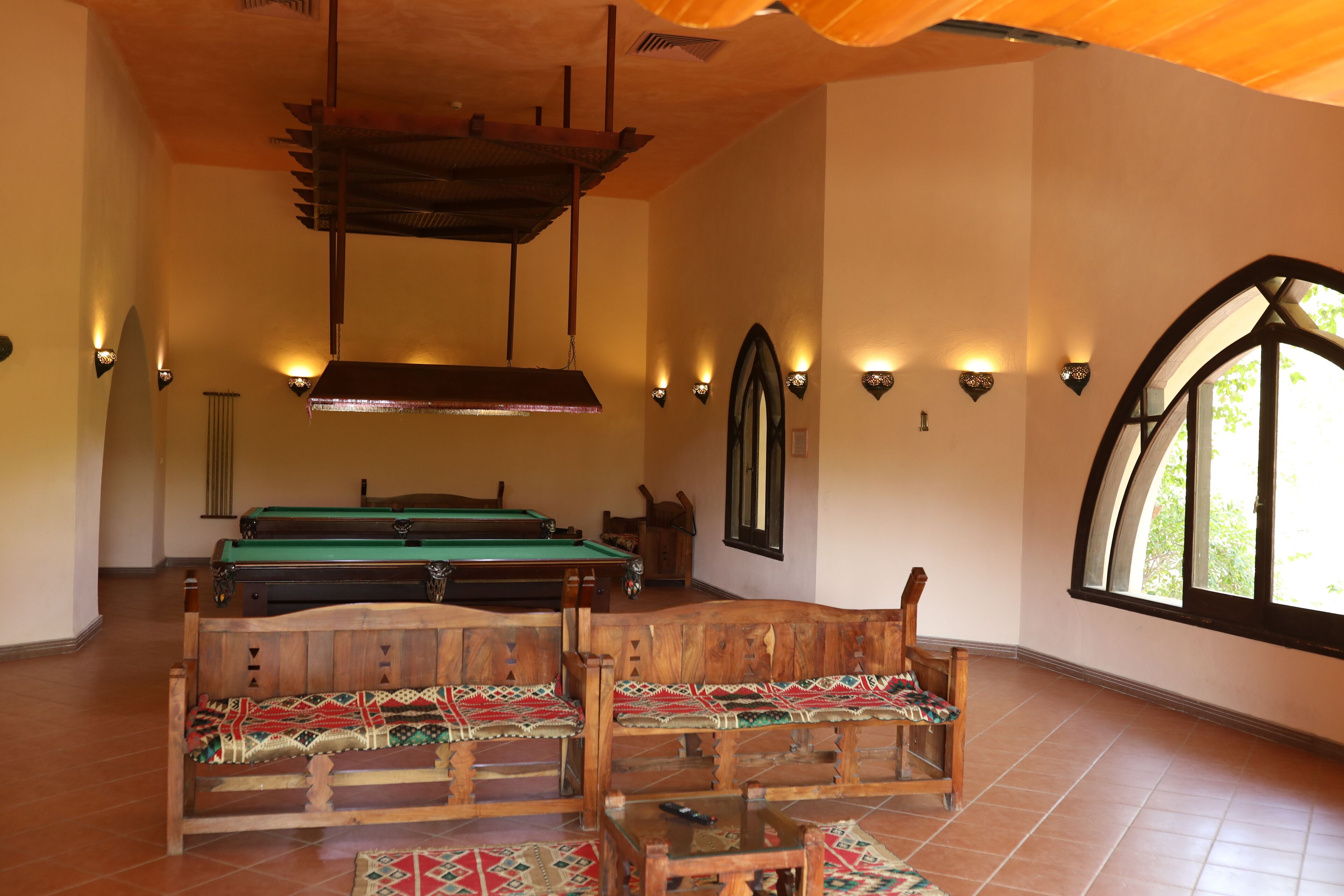 A pool table with a wooden bench in the foreground, a billiards table in the background, and a large arched window on the right.