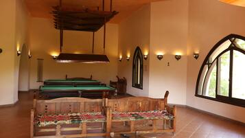 A pool table with a wooden bench in the foreground, a billiards table in the background, and a large arched window on the right.