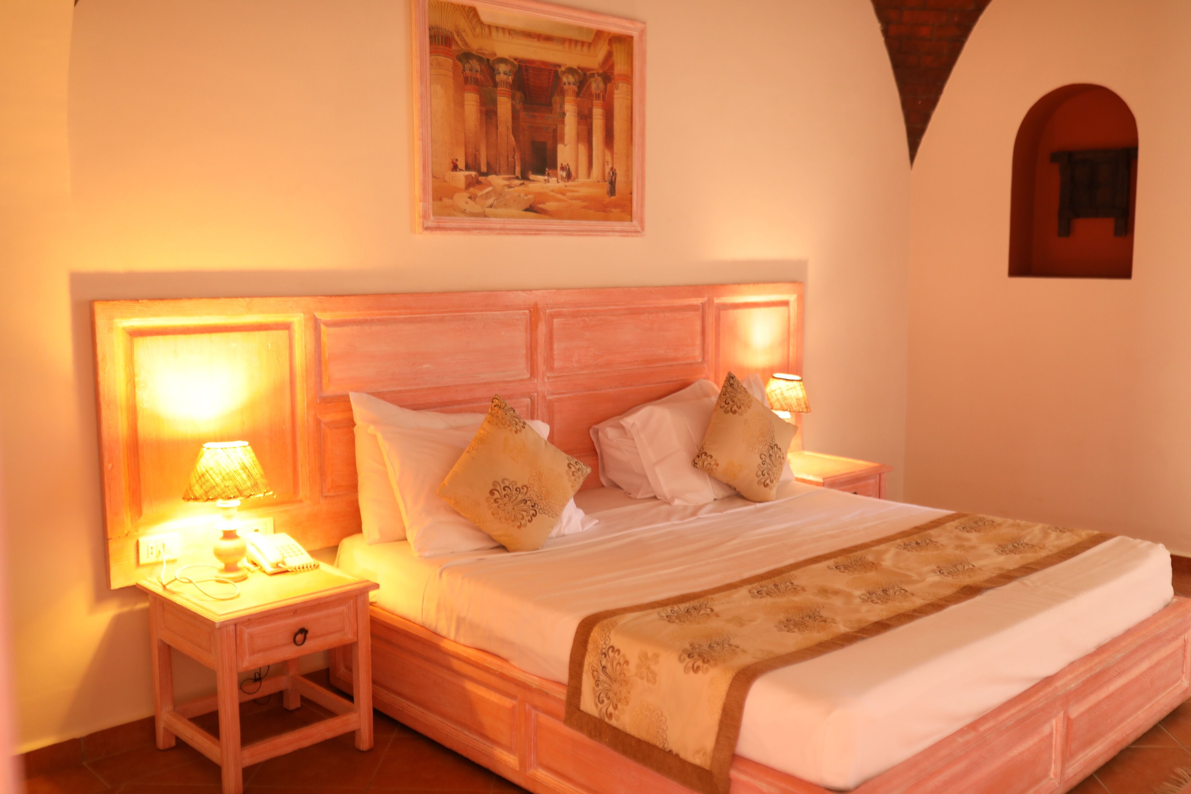 A bedroom with a wooden headboard, a bed with white linens and decorative pillows, a bedside lamp, and a painting above the bed.