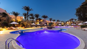 A swimming pool with a blue tint, surrounded by lounge chairs and umbrellas, palm trees, and buildings in the background. - TH Blue Lagoon Marsa Alam (Marsa Alam)