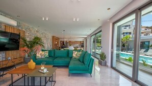 A modern living room with a green sectional sofa, a wooden coffee table, and a TV mounted on the wall. There is a large window offering a view of a swimming pool and buildings. - Glamorous Villa with Two Swimming Pools and great view (Chania)