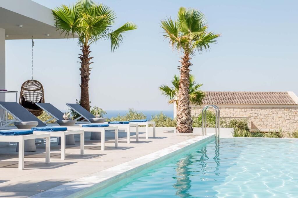 Un área junto a la piscina con sillones de descanso, palmeras y una vista despejada del mar.