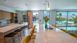 A modern kitchen with a long counter and bar seating, large windows with a view of greenery and mountains, and a bright interior. - Glamorous Villa with Two Swimming Pools and great view (Chania)