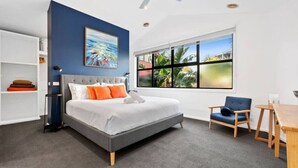 Un dormitorio moderno con cama gris, almohadas naranjas, pared azul y un ventanal con vistas a la vegetación.