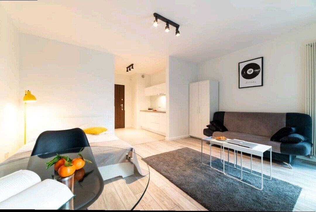 Un salon moderne avec une table à manger en verre, un canapé noir et une table basse. On aperçoit un coin cuisine au fond.