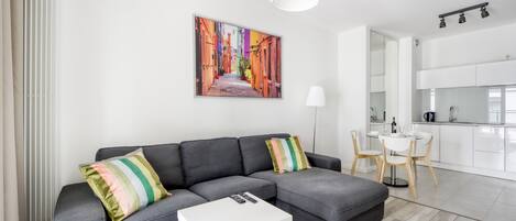 A modern living room with a grey sectional sofa, a white coffee table, and a colorful painting of a street scene. There is a dining area with a white table and chairs, and a kitchen with white cabinets and a pendant light.