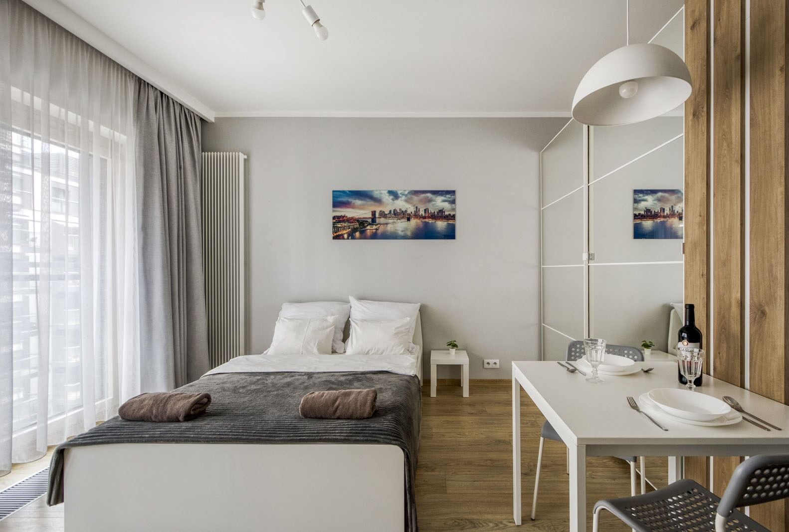 A modern hotel room with a bed, a dining table with chairs, a small side table, and a large window with curtains.