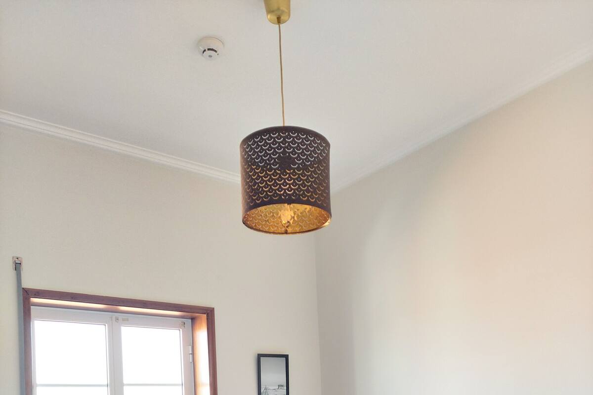 A bedroom with a bed, a window, a radiator, a nightstand, a lamp, and a hanging light fixture.