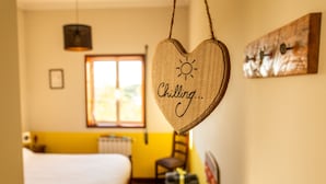 Ein Zimmer mit einem Bett, einem Fenster, einem herzförmigen Wanddekor mit dem Schriftzug 'Chilling...' und einem Sonnensymbol sowie einer Holzbildkunst.