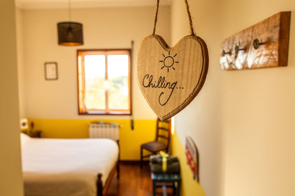 A room with a bed, a window, a hanging heart-shaped decoration with the word "Chilling..." and a sun symbol, and a wooden wall art piece.