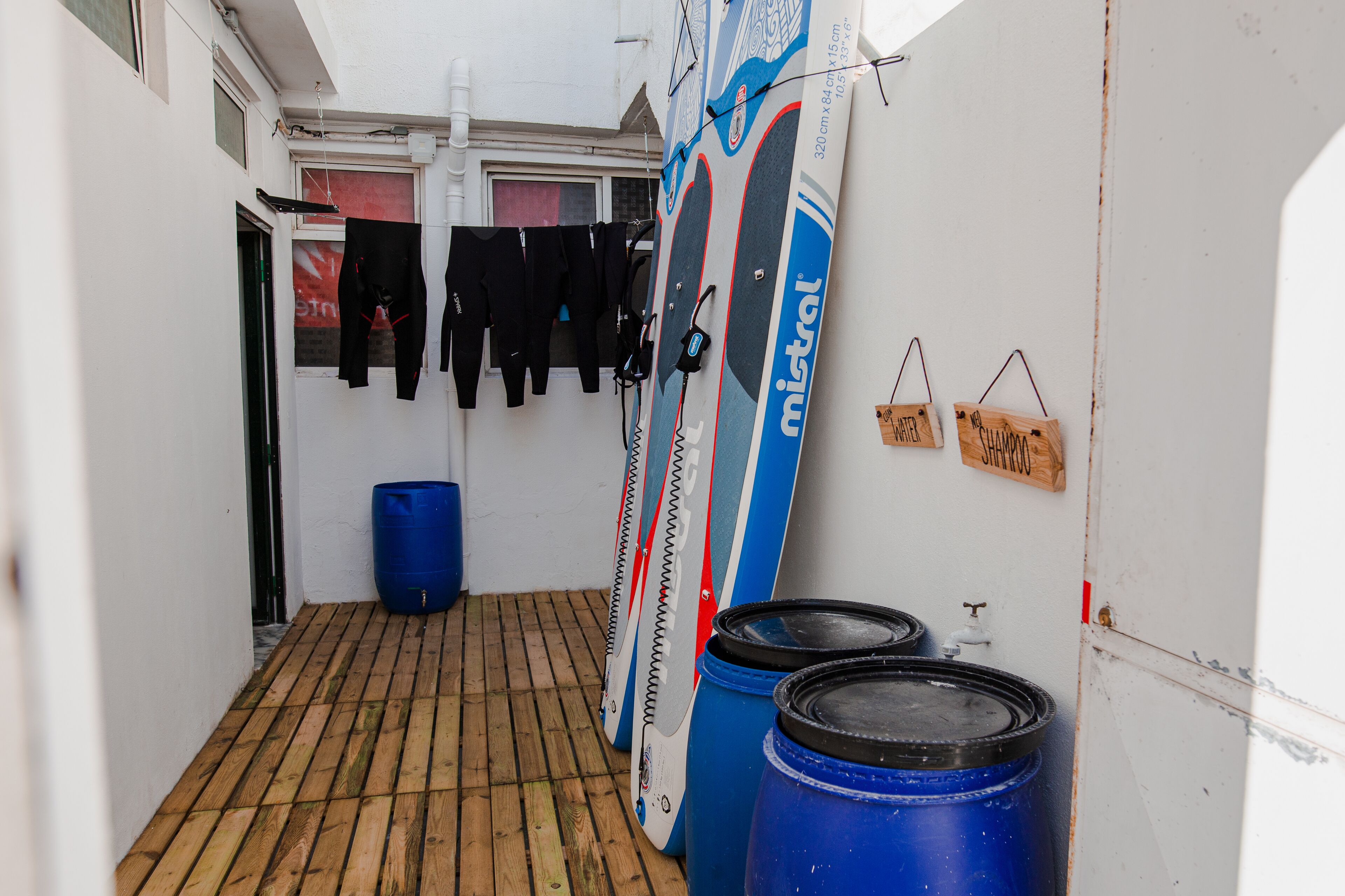 A narrow corridor with wooden flooring, a blue barrel, and surfboards leaning against the wall.