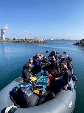 A group of people on a small inflatable boat with a lighthouse and rocky outcrop in the background. - My Way Kite & Surf House (Ilhavo)
