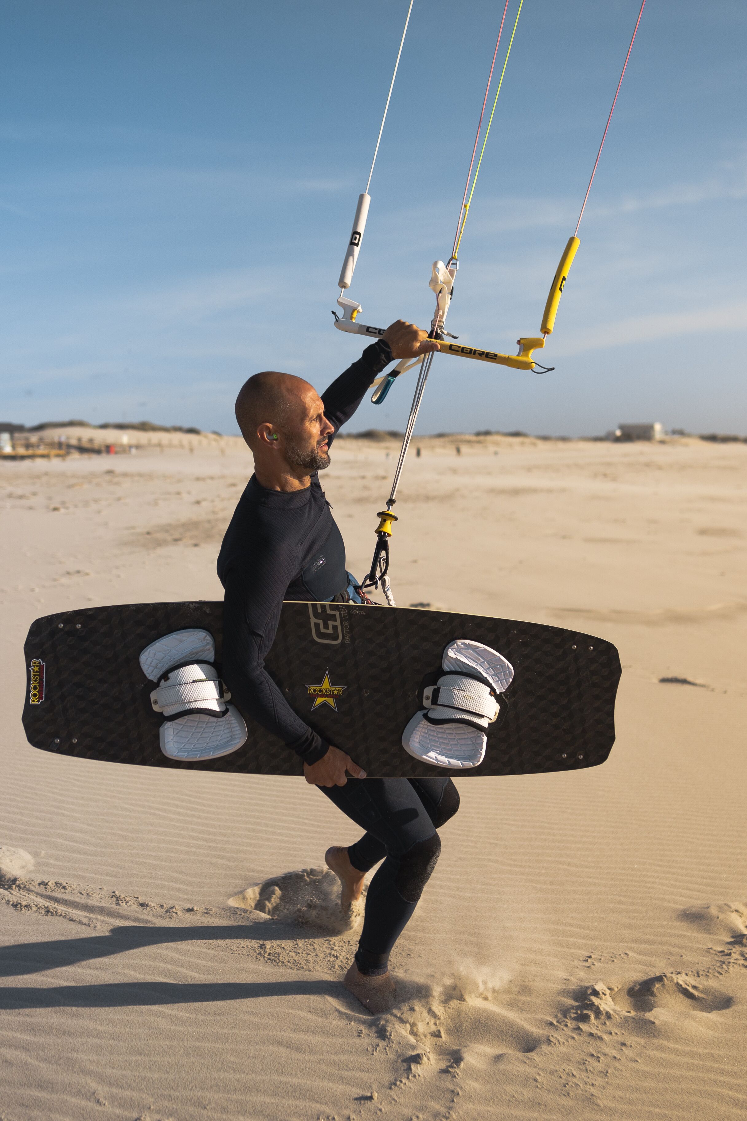 Una persona lleva una cometa para kitesurf grande con varias sujeciones y un asa, caminando sobre una playa arenosa.