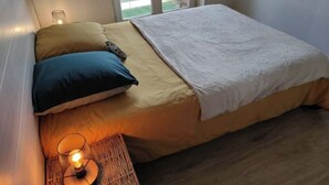Ein Einzelbett mit weißer Daunendecke, zwei Kissen (ein blaues, ein braunes), einer Nachttischlampe und einem gewebten Beistelltisch.