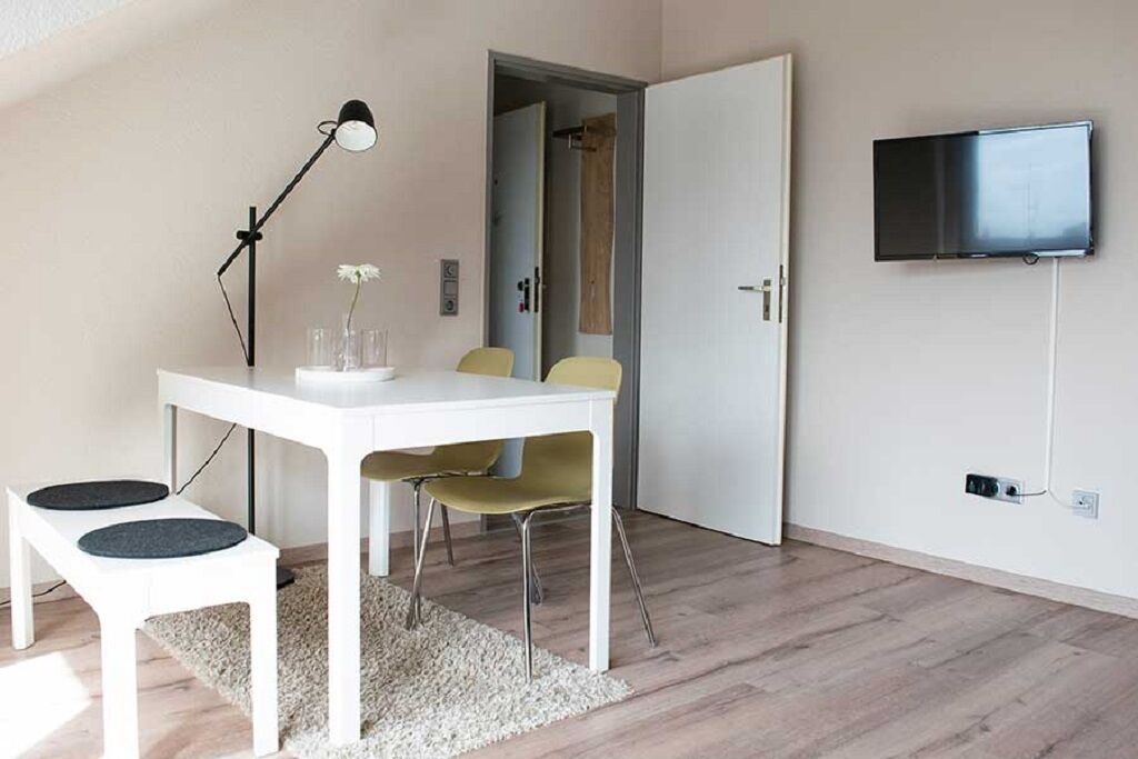 Et moderne værelse med et hvidt bord, to stole, en gulvlampe, et TV ophængt på væggen og en dør, der fører til et andet rum.