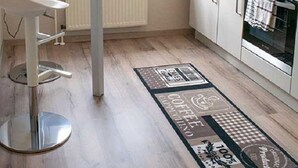 A compact kitchen with a white countertop, a small dining area with a white chair, a window with a view, and a patterned rug on the floor. - Schiller- Quartier 6 apartment "Heimathafen" (Bremerhaven)