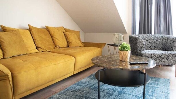 A living room with a yellow sofa, a patterned armchair, a round coffee table, and a small plant. - Schiller- Quartier 6 apartment "Heimathafen" (Bremerhaven)